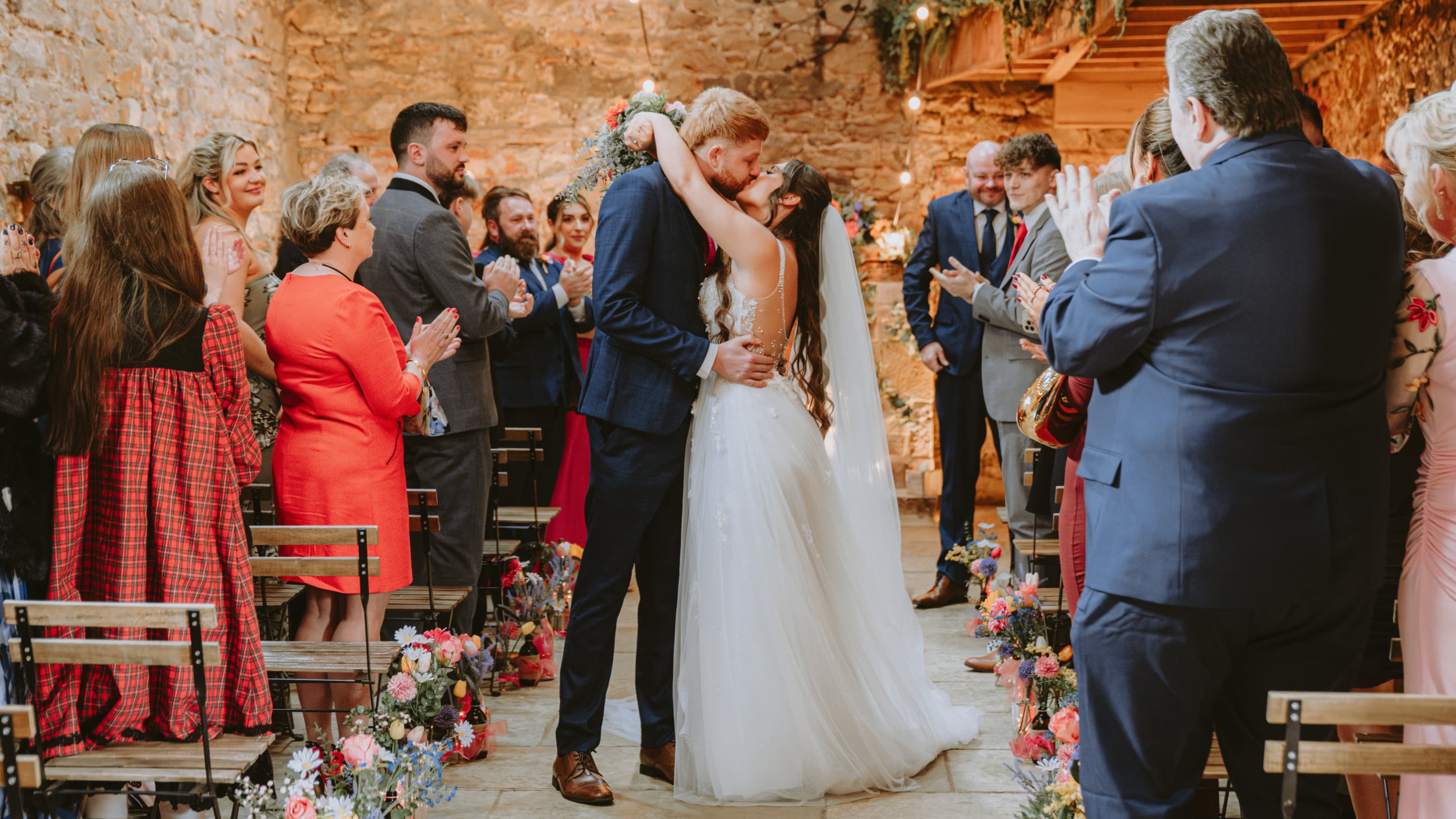 Bride and groom kiss in the aisle during a rustic indoor wedding ceremony, surrounded by applauding guests, with floral arrangements lining the aisle and warm string lighting overhead.
